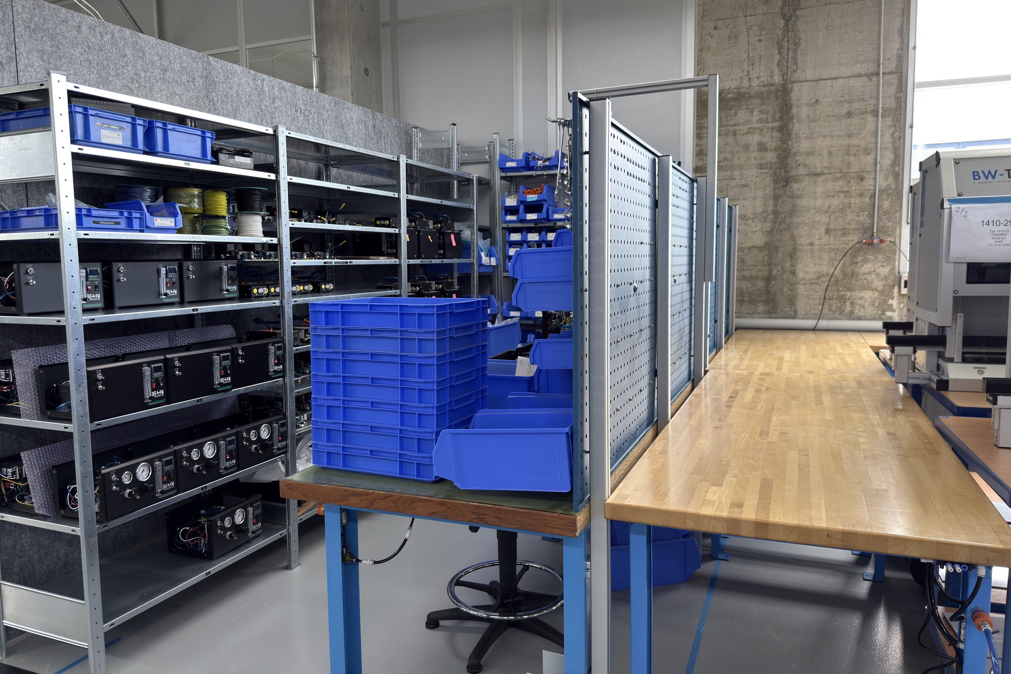 Photo of a metal rack with equipment boxes, blue bins and a wooden workbench with a machine.