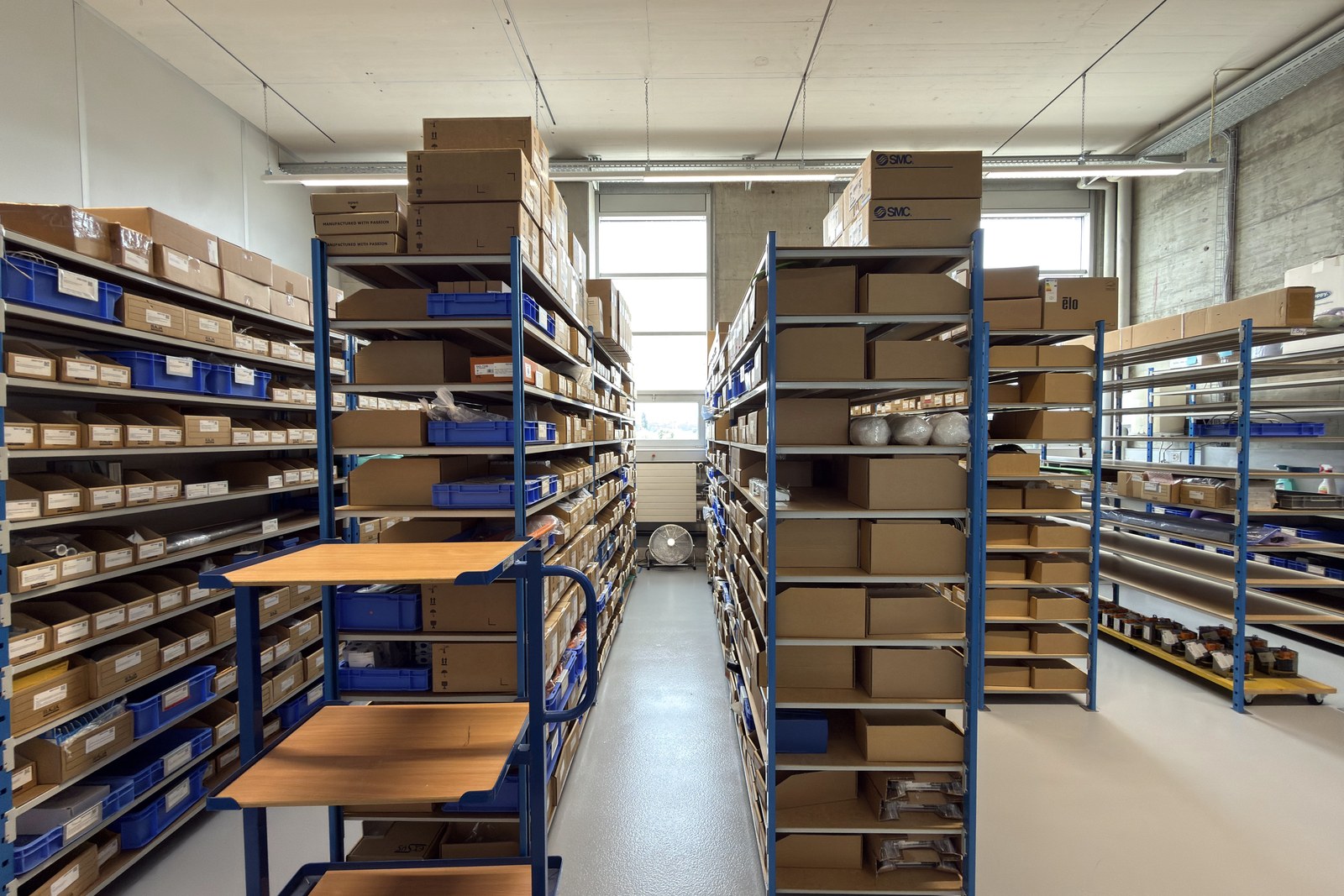 Aisle between blue steel shelving racks filled with boxes and bins, under a light ceiling with tall windows at the end.