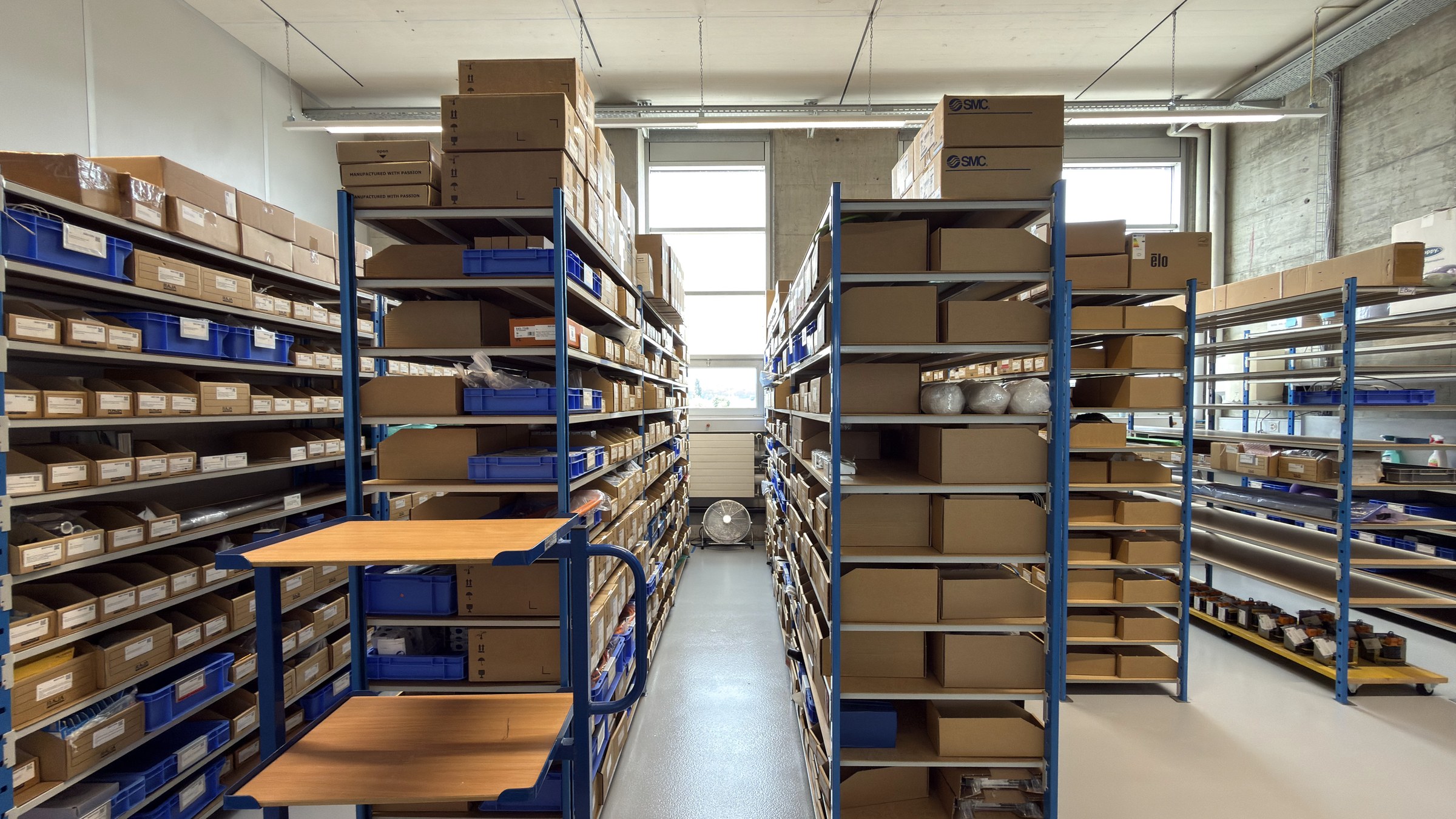 Aisle between blue steel shelving racks filled with boxes and bins, with a window at the end of the hall.