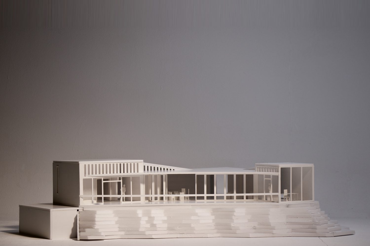 White model of a long glazed pavilion with a slatted roof edge, standing on a base of stacked stepped layers.