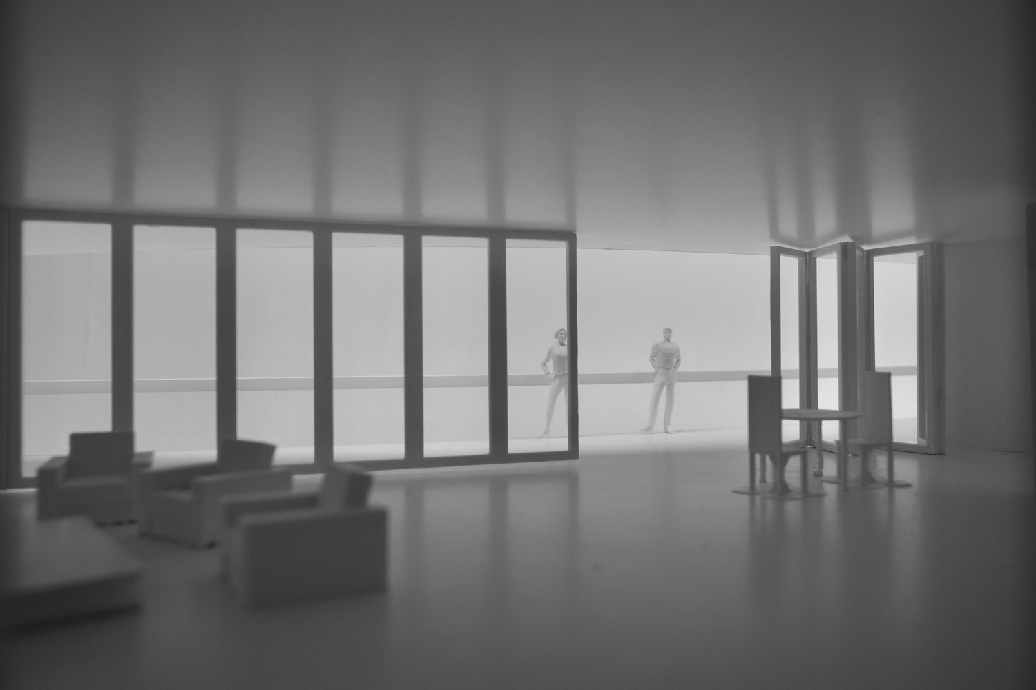Black and white photo inside the pavilion model: armchairs in front, a glazed partition, a table with chairs and two standing figures.