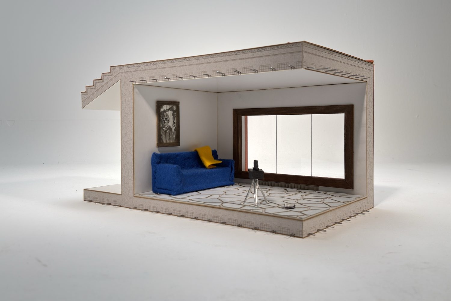 Model of a room cut open: a blue sofa with a yellow throw, a framed picture, a large window and a camera on a tripod on a floor of irregular paving.