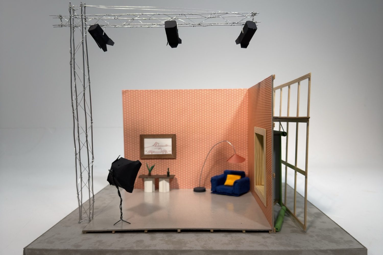 Model of a room with a brick wall, an armchair and an arched lamp, lit by a wire truss with black stage lights.