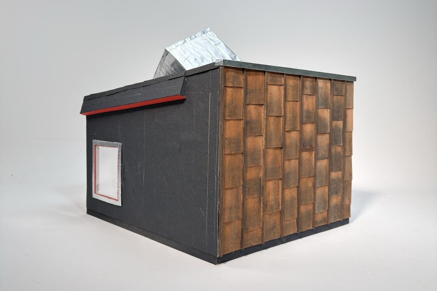 Model of a box with a black wall and window on one side, timber shingles on the other and a foil-clad roof element on top.