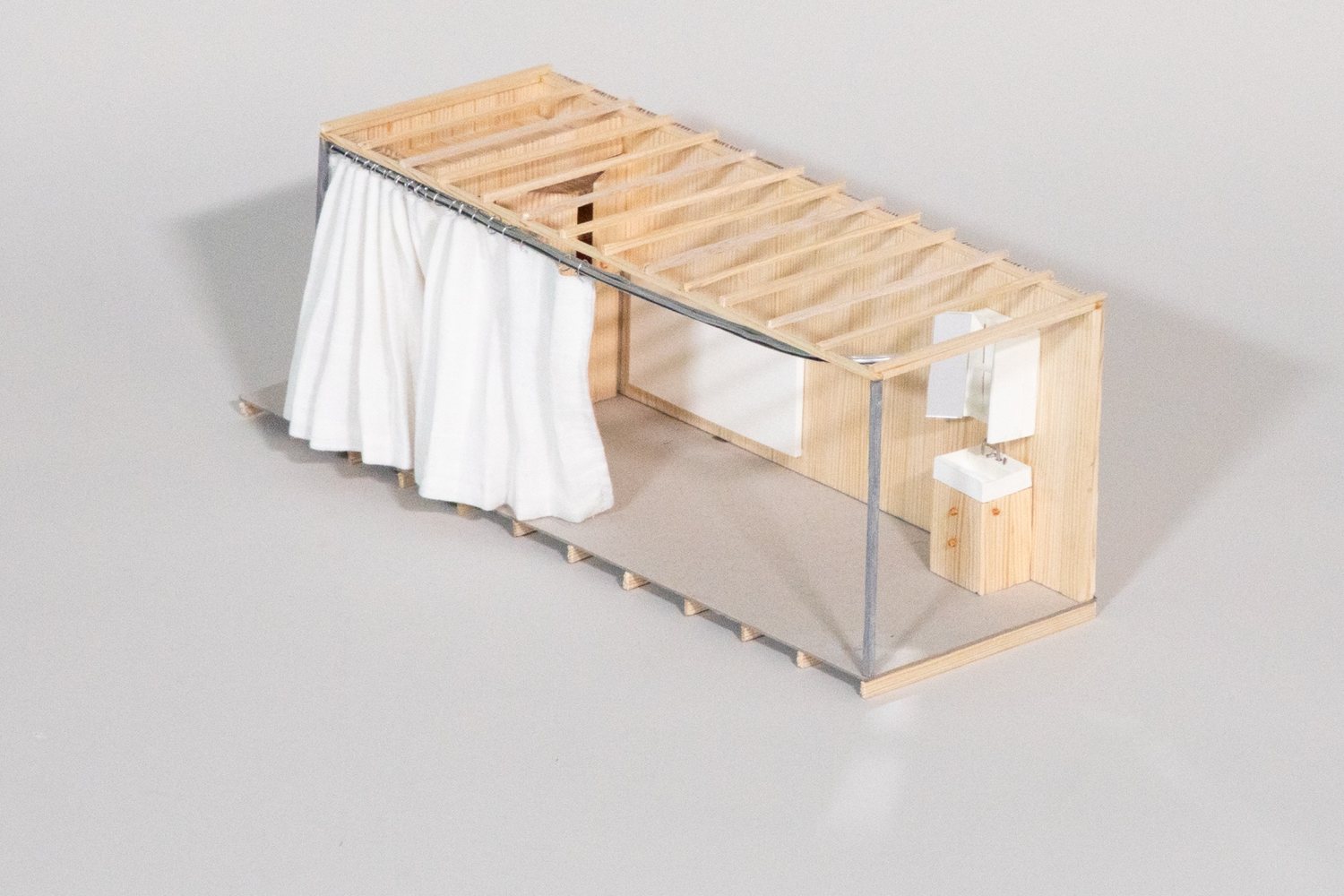 Timber model of a small room with a slatted roof, a white curtain along one side and a washbasin at the end.