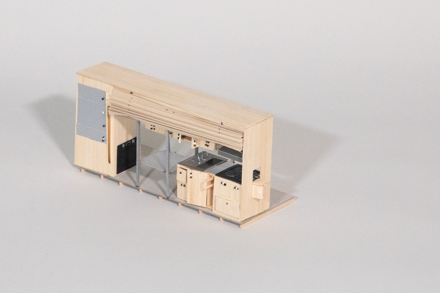 Timber model of a kitchen module: cupboards, a sink and a cooktop under a timber canopy, with an oven and a grey cabinet on the left.