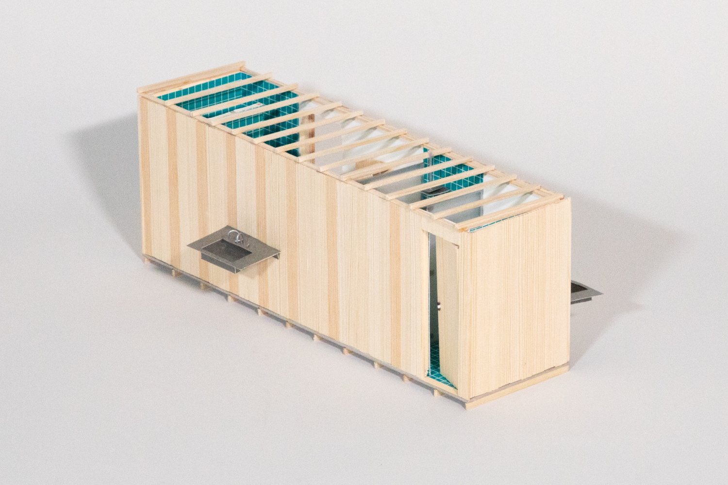 Timber model of a long narrow room clad in vertical boards, with a washbasin on the outer wall, a door at the end and teal tiled surfaces visible through the open roof.