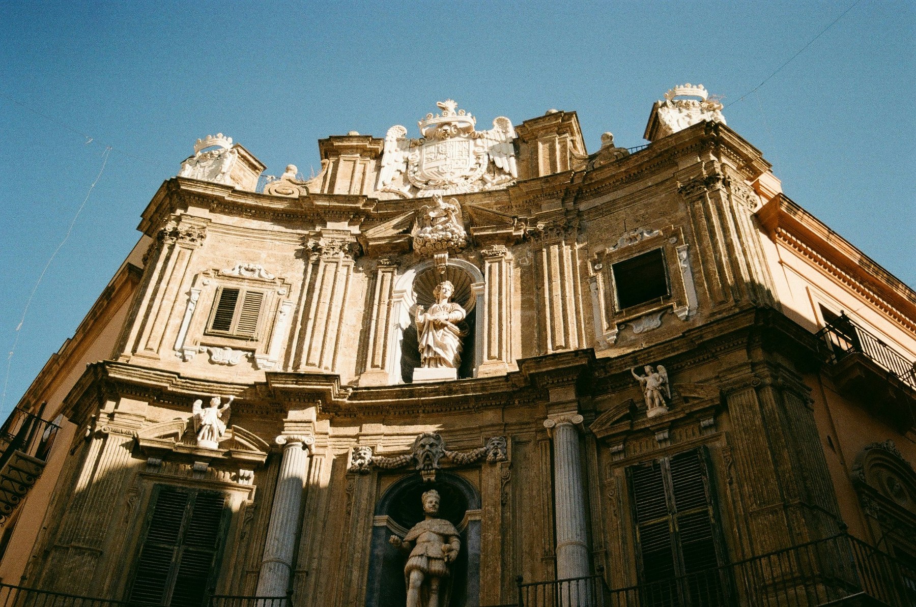 Low-angle view of a baroque corner facade with statues, columns and a coat of arms against a blue sky, shot on film.