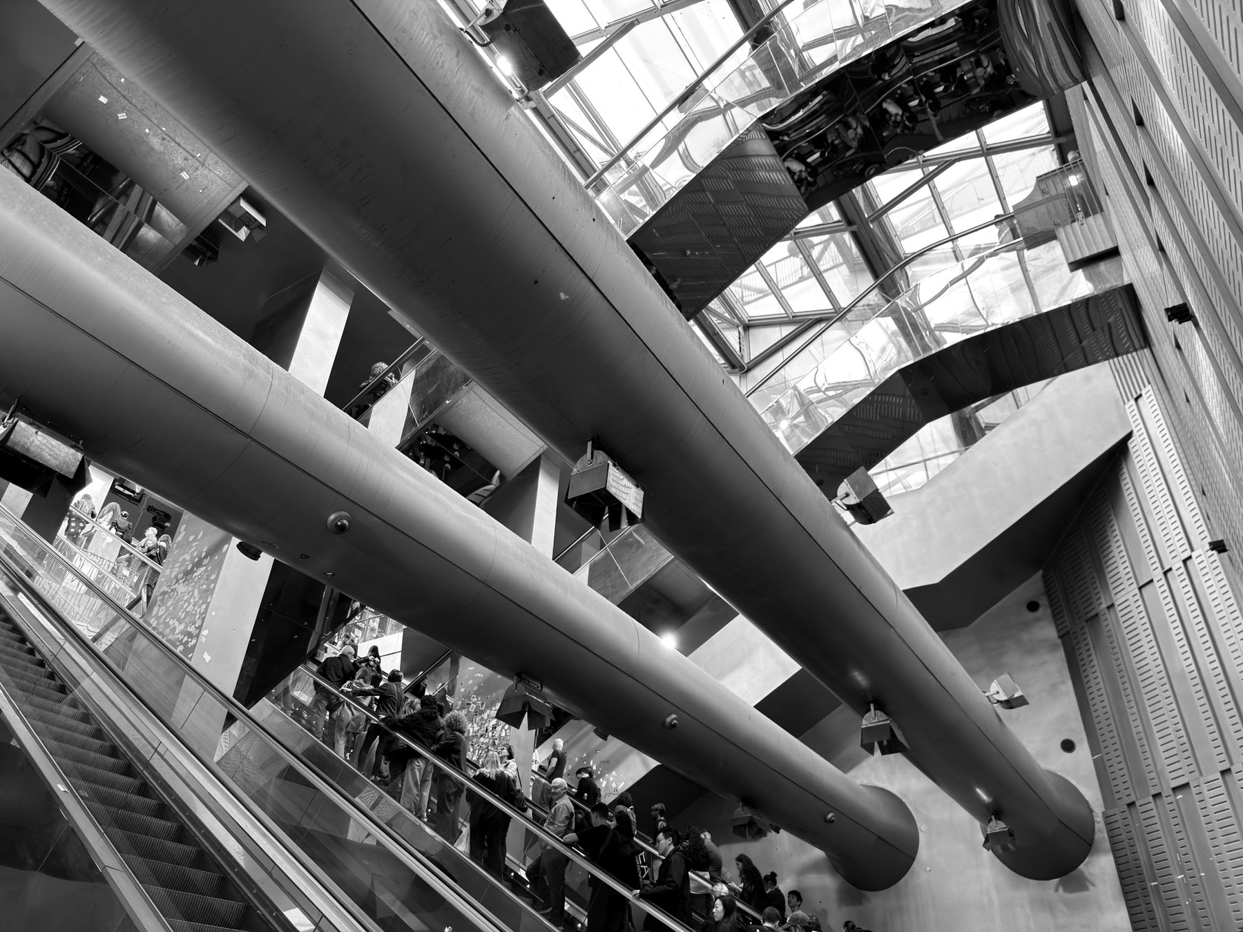 Black and white view of large steel tubes crossing a station hall above escalators with people, with mirrored cladding and a glazed roof.