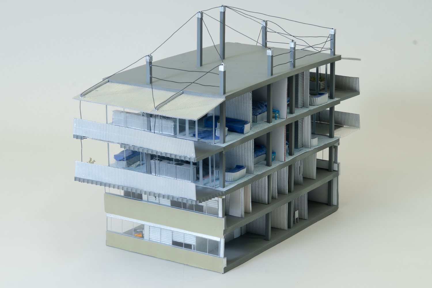 Grey card model of the building seen from a corner: balconies with corrugated screens, an awning at the top and posts with cables on the roof.